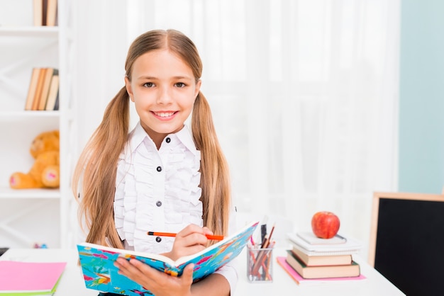 Free photo smiling schoolgirl writing task with pencil in notebook at classroom