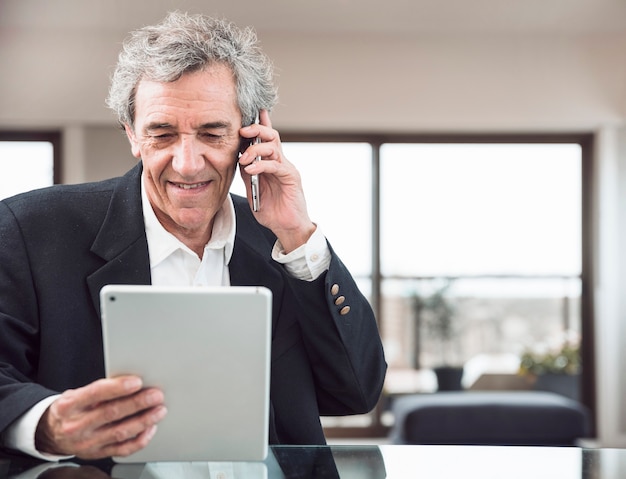 Smiling senior man talking on mobile phone looking at digital tablet at workplace