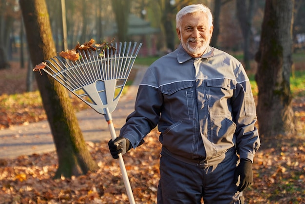 Smiling senior worker posing to camera while gathering autumn leaves outdoors portrait of happy