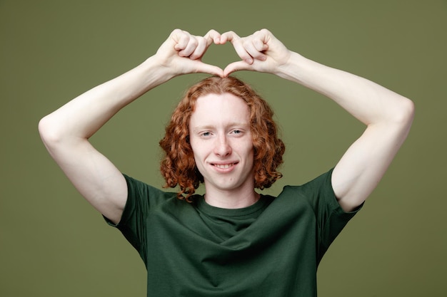 Free photo smiling showing heart gesture young handsome guy wearing green t shirt isolated on green background