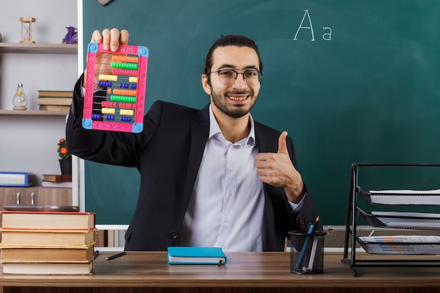 Smiling showing thumb up male teacher wearing glasses holding abacus sitting at table with school tools in classroom