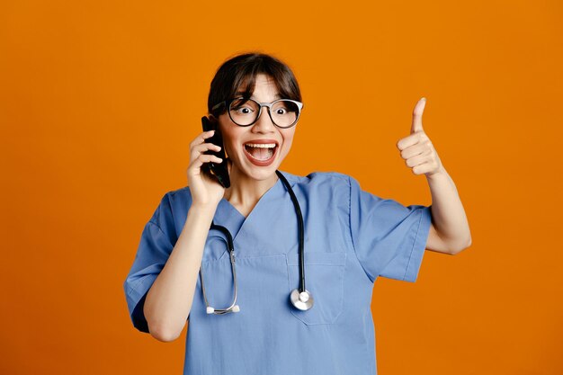 Smiling showing thumbs up speaks on the phone young female doctor wearing uniform fith stethoscope isolated on orange background