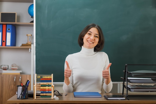 smiling showing thumbs up young female teacher sitting at desk with school tools on in classroom