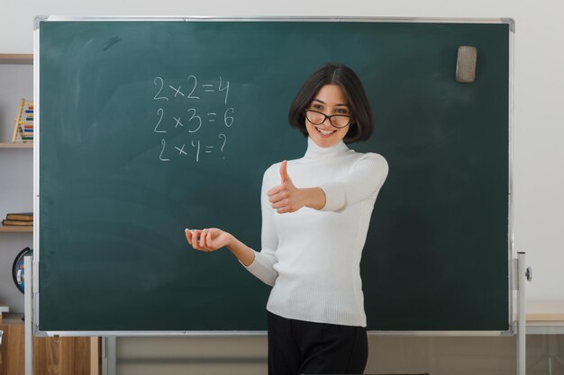 smiling showing thumbs up young female teacher wearing glasses standing in front blackboard and writes in classroom