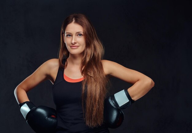 Smiling sportive woman dressed in sportswear wearing boxing gloves posing in a studio. Isolated on a dark textured background.