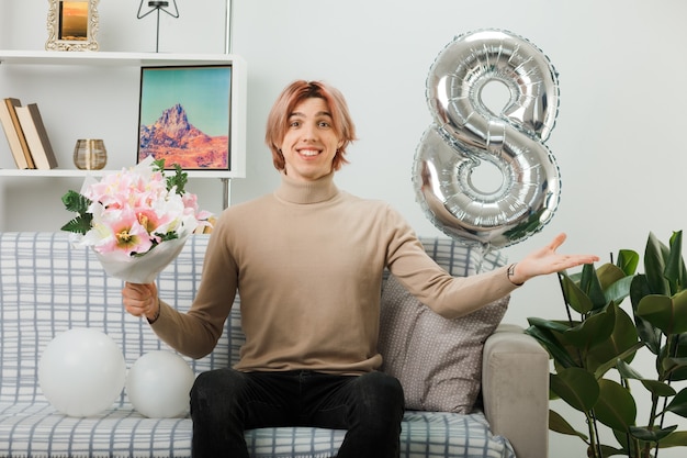 Smiling spreading hands handsome guy on happy women day holding bouquet sitting on sofa in living room