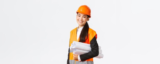 Smiling successful asian female architect construction engineer in safety helmet carry blueprints an