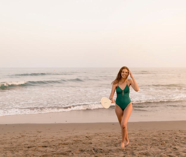 Smiling teenage girl in green bikini holding racket standing near the seashore