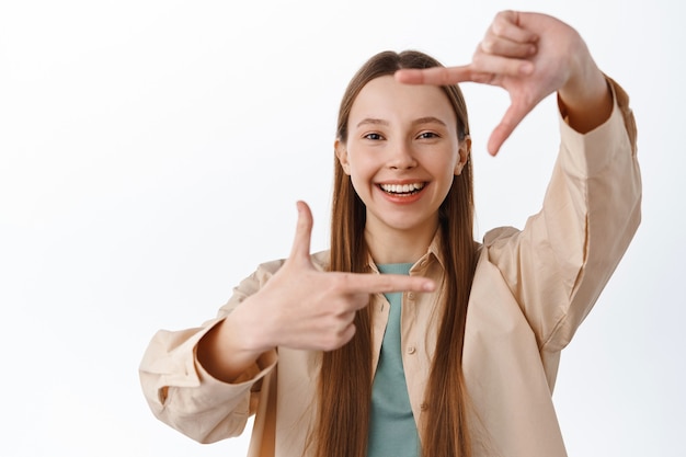 Smiling teenage girl look through hand frames camera gesture, picturing something, measure perfect angle, standing over white wall.