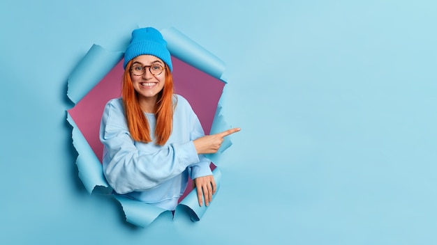 Smiling teenage girl with red hair gives recommendation, pointing at copy space, breaks through blue paper hole