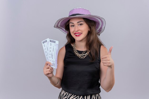 Smiling traveler young girl wearing black undershirt in hat holding suitcase and her thumb up on white background