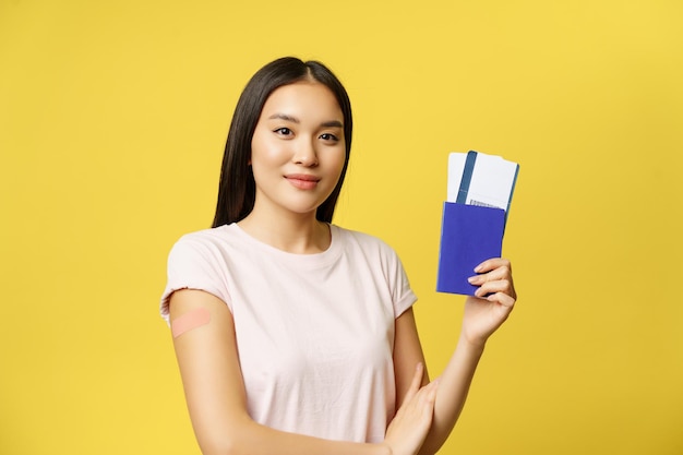 Smiling vaccinated from covid asian girl, showing passport with tickets, getting coronavirus vaccine shot for travelling abroad, yellow background