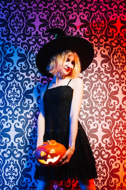 Free Photo smiling witch holding pumpkin by wall