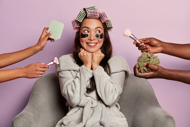 Free Photo smiling woman applies eye patches and hair curlers, surrounded by many beauticians