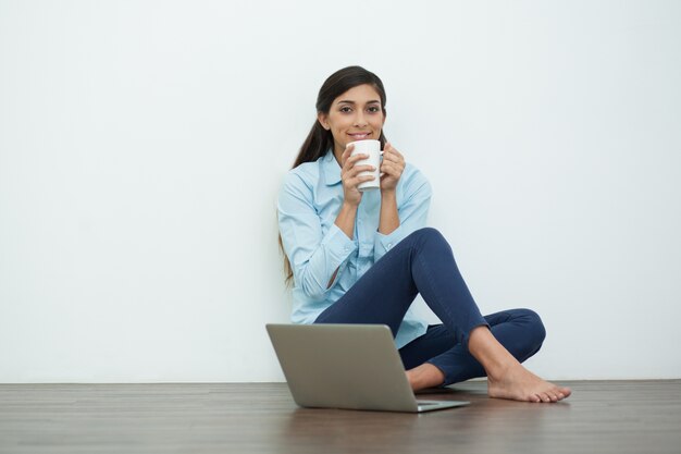 Smiling Woman Drinking Tea on Floor with Laptop