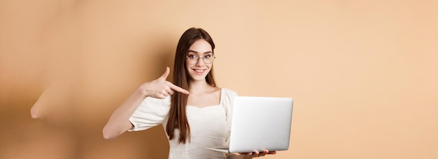 Free Photo smiling woman in glasses pointing finger at laptop screen showing online promo standing on beige bac