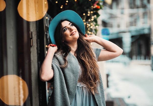 Smiling woman in hat and sweater stands in light dress on a winter street 