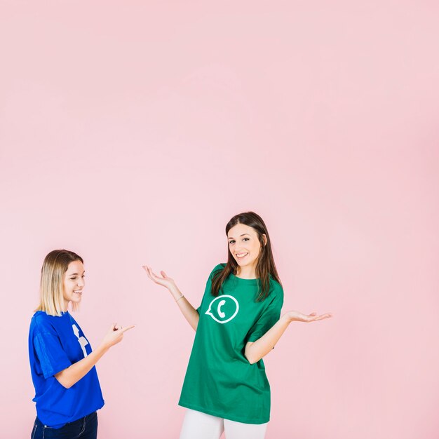 Smiling woman pointing at her friend shrugging on pink background