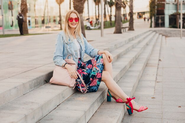 smiling woman sitting on stairs in city street in stylish printed skirt and denim oversize jacket with leather backpack wearing pink sunglasses