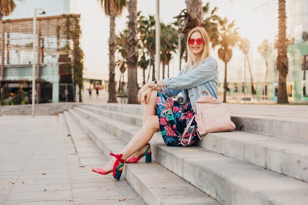 smiling woman sitting on stairs in city street in stylish printed skirt and denim oversize jacket with leather backpack wearing pink sunglasses