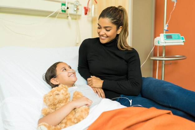 Smiling woman talking to sick daughter while sitting on bed at hospital