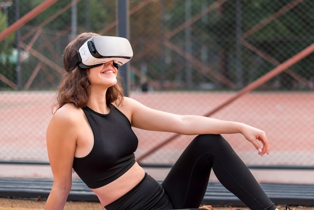 Free photo smiling woman in vr headset in a tracksuit sitting on yoga mat on a sports field in a park