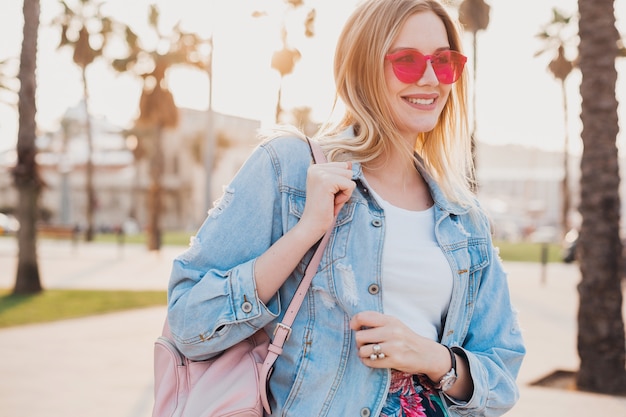 Free Photo smiling woman walking in city street in stylish denim oversize jacket wearing pink sunglasses, holding leather backpack