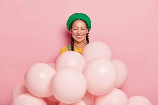 Smiling woman with cheerful expression, keeps eyes closed from pleasure, wears green beret, stands with inflated helium balloons