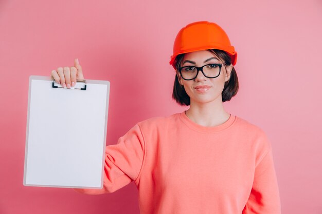 Free Photo smiling woman worker builder hold white sign board blank against pink background. building helmet.