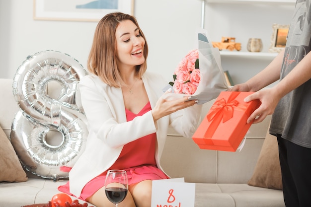 Smiling women's sitting on sofa on happy women's day gives present with bouquet by someone in living room