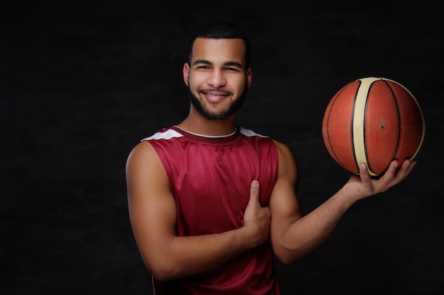 Smiling young African-American basketball player in sportswear over dark background.