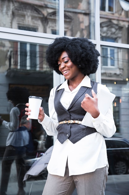 Smiling young african businesswoman holding takeaway coffee cup and digital tablet in hand