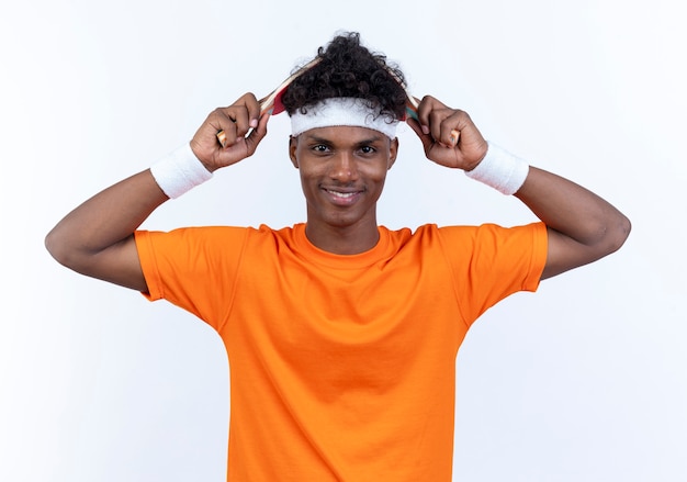 Smiling young afro-american sporty man wearing headband and wristband covered head with ping pong racket isolated on white wall