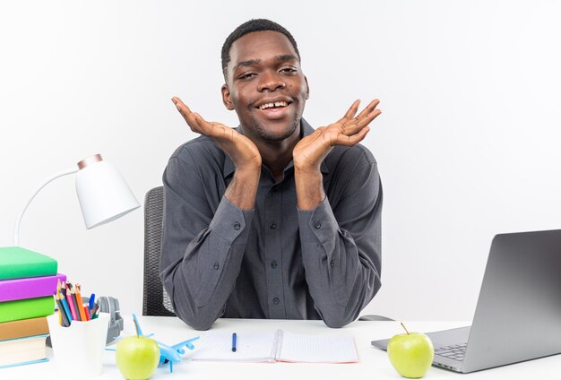 Smiling young afro-american student sitting at desk with school tools keeping hands open