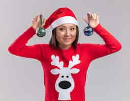 Free photo smiling young asian girl wearing christmas hat with sweater holding christmas tree balls isolated on white background