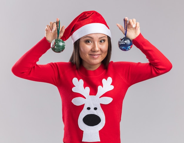 Free Photo smiling young asian girl wearing christmas hat with sweater holding christmas tree balls isolated on white background