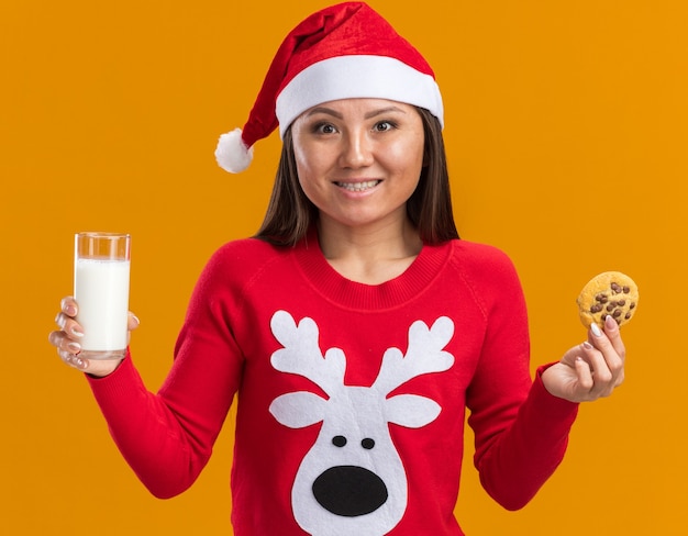 Free photo smiling young asian girl wearing christmas hat with sweater holding glass of milk with cookies isolated on orange wall