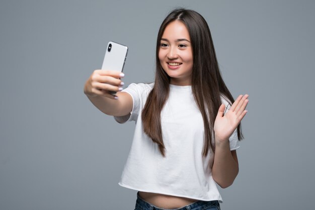 Smiling young asian woman taking a selfie with mobile phone over isolated gray wall background