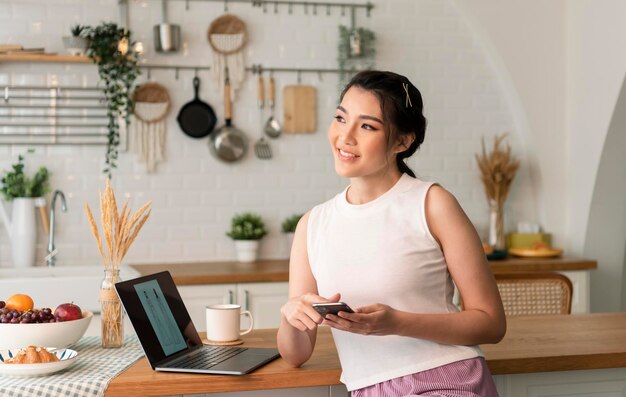 Smiling young asian woman using mobile phone while sitting in kitchen room at home with laptop