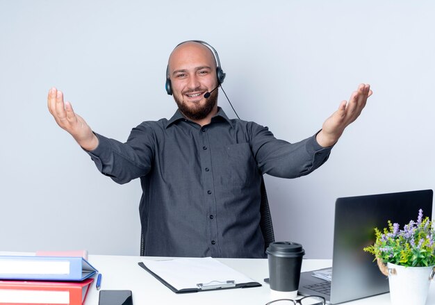 Smiling young bald call center man wearing headset sitting at desk with work tools showing empty hands isolated on white wall