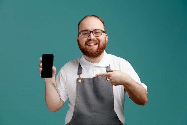 Smiling young barber wearing uniform and glasses looking at camera showing mobile phone pointing at it isolated on blue background