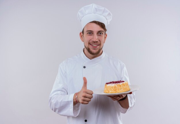 A smiling young bearded chef man wearing white cooker uniform and hat holding a plate with cake and showing thumbs up while looking on a white wall