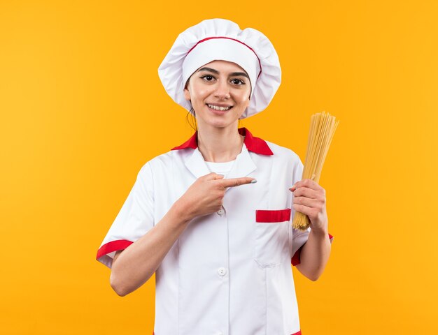 Smiling young beautiful girl in chef uniform holding and points at spaghetti 