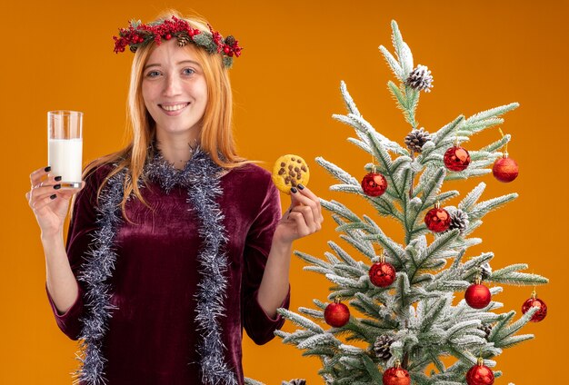 smiling young beautiful girl standing nearby christmas tree wearing red dress and wreath with garland on neck holding glass of milk with cookies isolated on orange wall