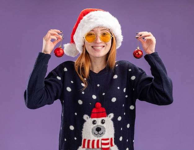 Free photo smiling young beautiful girl wearing christmas sweater and hat with glasses holding christmas balls isolated on purple background