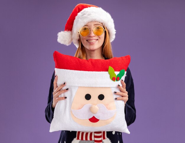 Smiling young beautiful girl wearing christmas sweater and hat with glasses holding christmas pillow isolated on purple background