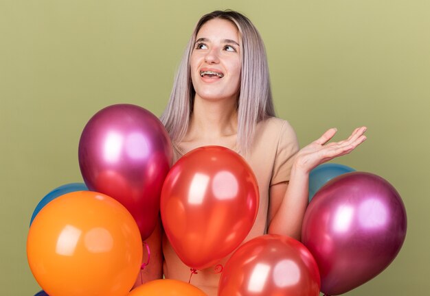 Smiling young beautiful girl wearing dental braces standing behind balloons spreading hand isolated on olive green wall