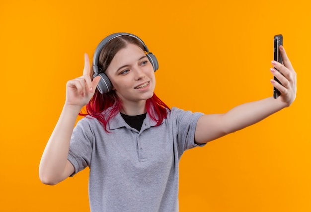 Free Photo smiling young beautiful girl wearing gray t-shirt in headphones points finger to up taking selfie on isolated yellow background