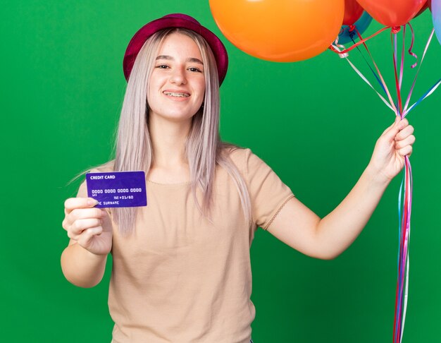 Smiling young beautiful girl wearing party hat and braces holding balloons with credit card 