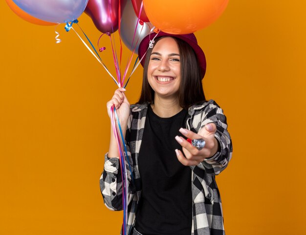 Smiling young beautiful girl wearing party hat holding balloons and holding out hand at camera 